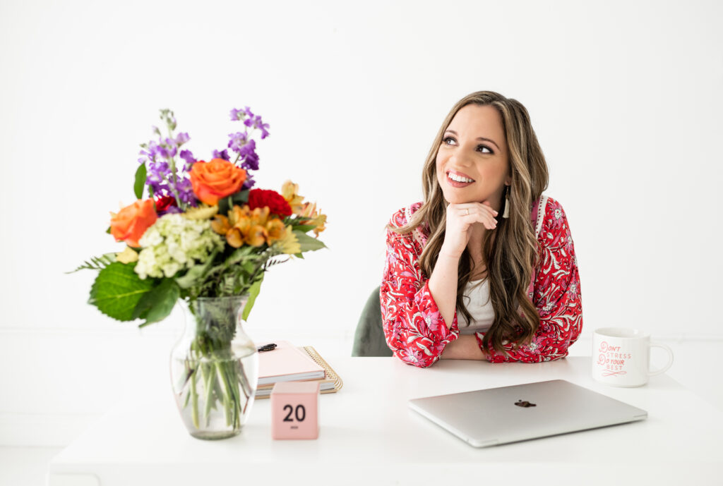 a woman is seated at a white desk with a cheerful, inviting smile, looking slightly upward. On the desk in front of her is a vase filled with vibrant flowers, a pink calendar block displaying the 20th, a laptop, a notebook, and a coffee mug that says, "Don’t stress, do your best." | how i use enji to market my wedding business