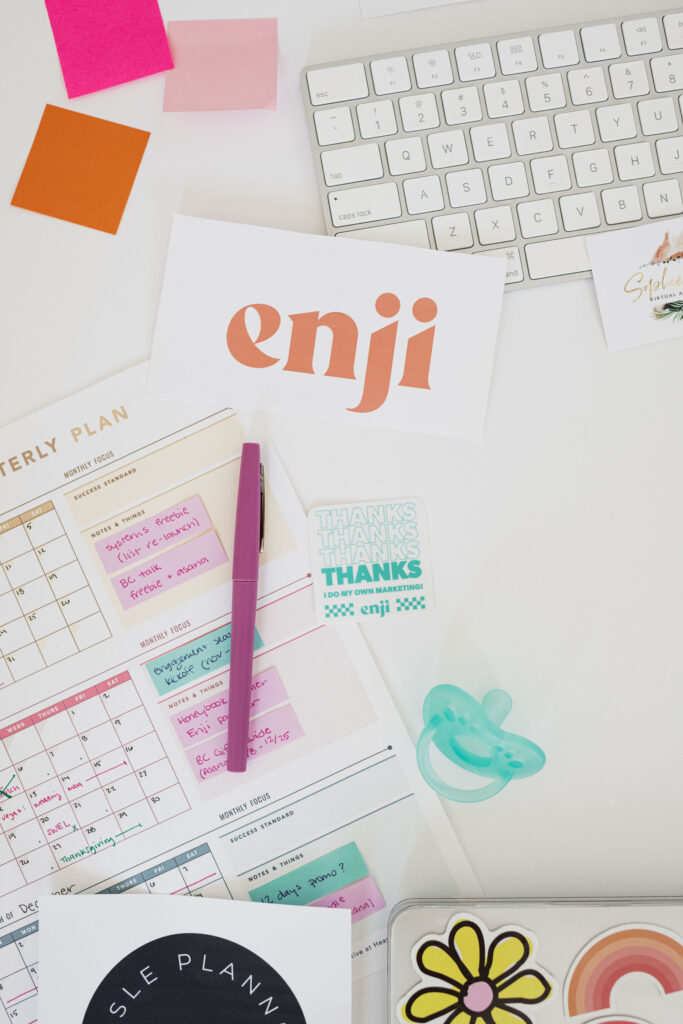 A bright, organized workspace featuring a keyboard, colorful sticky notes, a planner with notes, a purple pen, an "enji" card, a pacifier, and playful stickers.